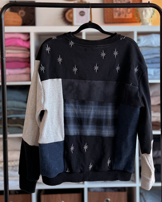 Black, blue and tan patchwork sweater with star patterns and plaid design on a rack