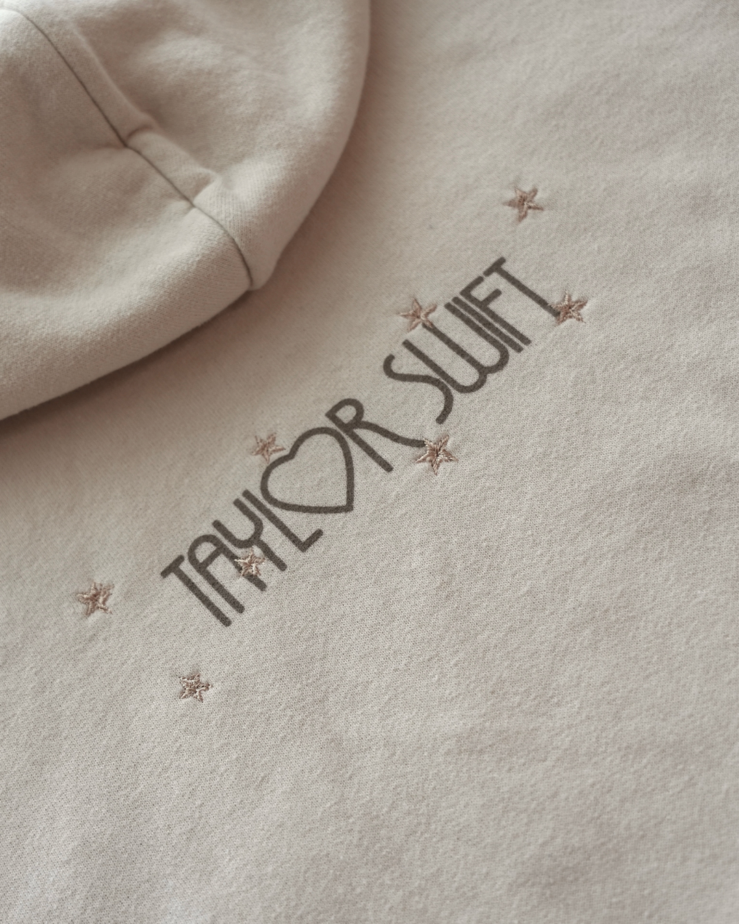 Beige sweatshirt with 'Taylor Swift' text and embroidered star designs on a soft focus background